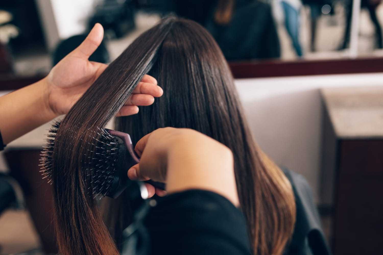 Stylist brushing and straightening a woman's long, smooth hair in a salon.