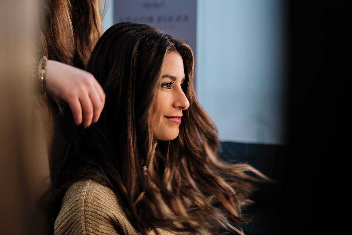 Woman smiling as stylist adjusts her long, wavy hair indoors.
